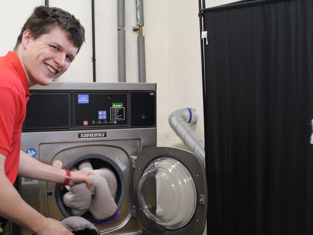 Elliot one of our volunteers posing in a red polo t-shirt, putting a grey dog toy in a grey washing machine.