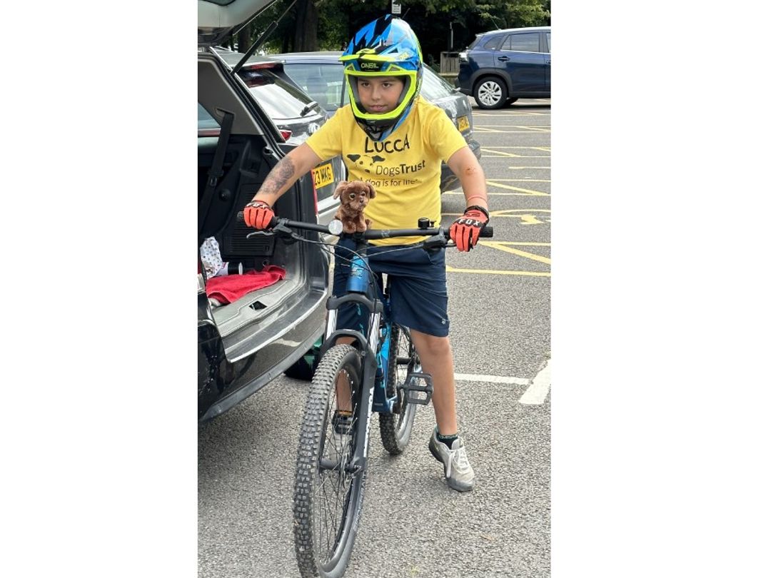 Lucca an eight-year-old dog lover from Doncaster, smiling on a bike with a blue helmet next to black car in a car park. 