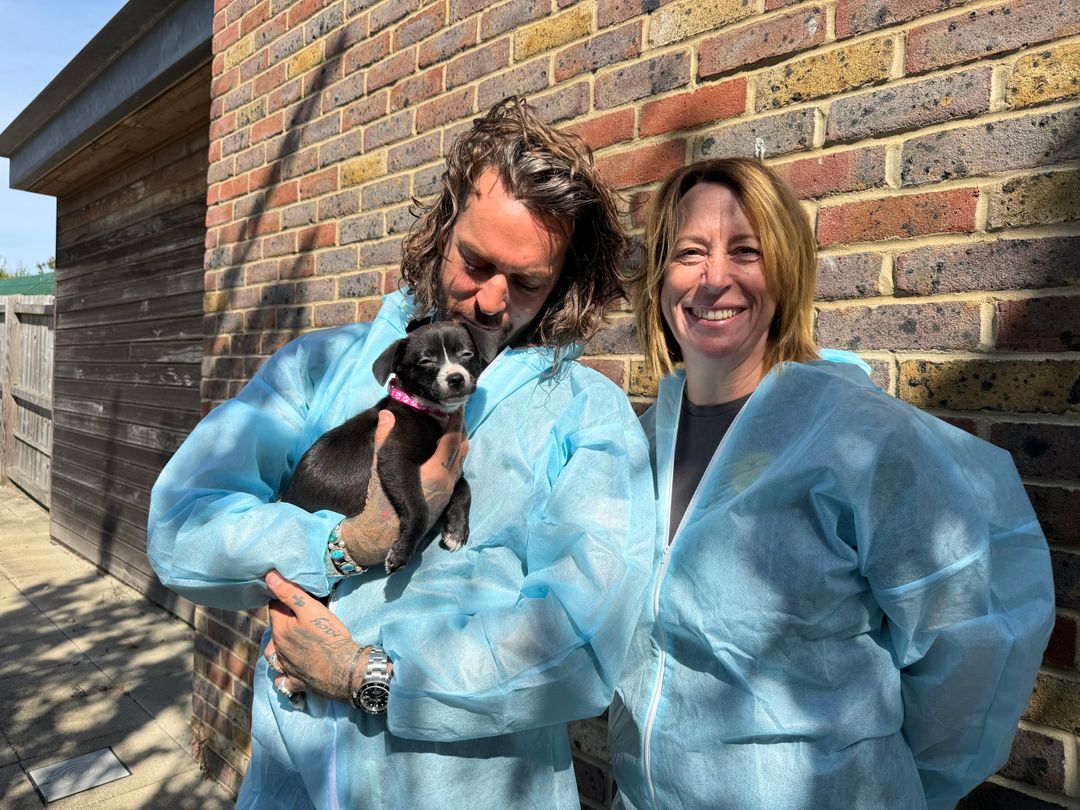Pete wickes standing next to Basildon centre manager Lisa with Pete holding a black puppy. They are both wearing blue PPE boiler suits