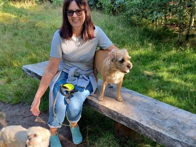 a lady smiling wearing sunglasses sat next to two beige small dogs on a bench