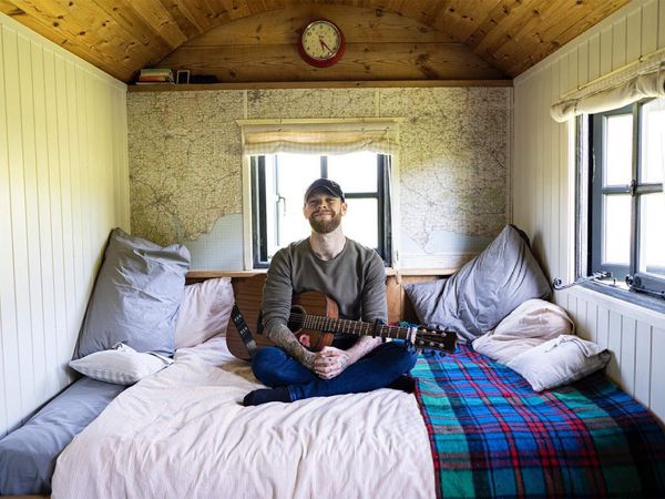 Frazer Lepford, A Cheltenham-based singer-song writer who was Amazon’s Breakthrough Artist of 2021. Sitting and smiling cross-legged in a bedroom with pillows and checkered blanket.