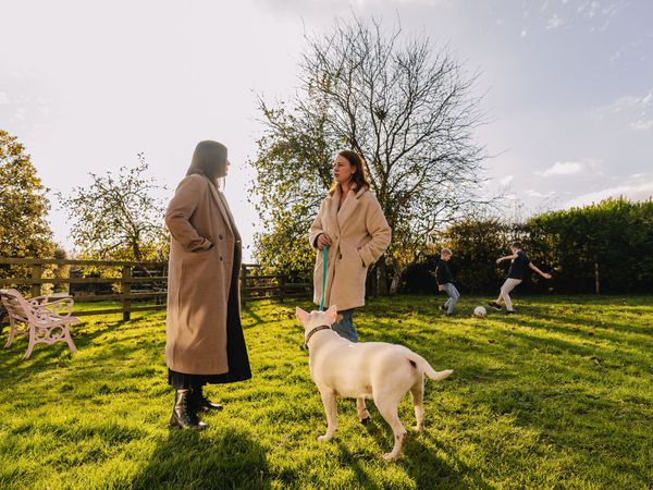 A white bull terrier standing next to two ladies chatting in a park, whilst two boys play football in the background