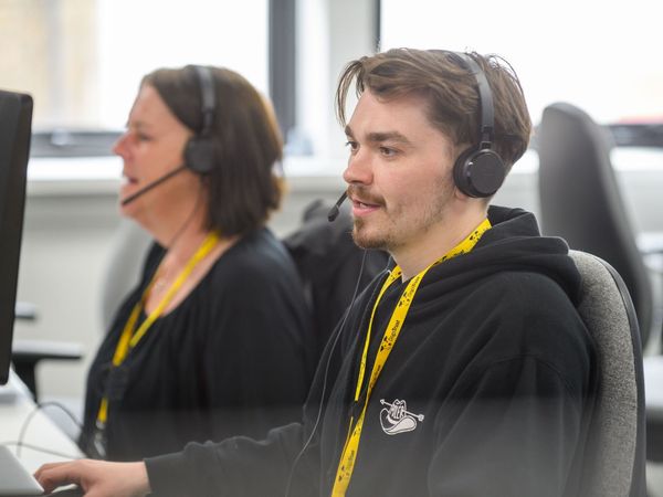 male staff member speaking to someone via telephone head set, sat at a computer at a desk