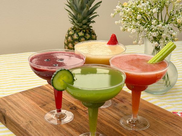 Four colourful smoothies in cocktail glasses on a wooden board, each with a different garnish.