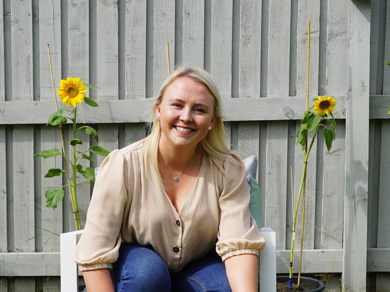 Polly a Dogs Trust foster carer, sitting on a white chair, smiling in a lovely green garden with sunflowers in the background.