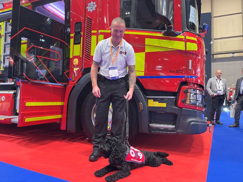 Alan Gale firefighter behind a firetruck, with OK9 black cocker spaniel cross dog in ok9 vest laying on the floor.