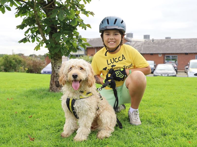 Lucca an eight-year-old dog lover from Doncaster, smiling in a green park bending down next to a beige Cockapoo dog. Lucca has a yellow t-shirt one and a black helmet.  