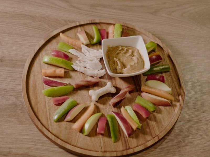 Our charcuterie-style board for dogs. Circular wooden board with a small bowl of peanut butter, treats, chicken, and sliced apples, green and red bell peppers and carrot slices displayed in a circle.