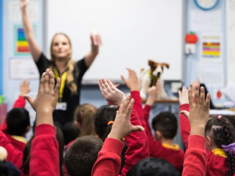 Education events teacher in classroom with lots of children holding their hands up.