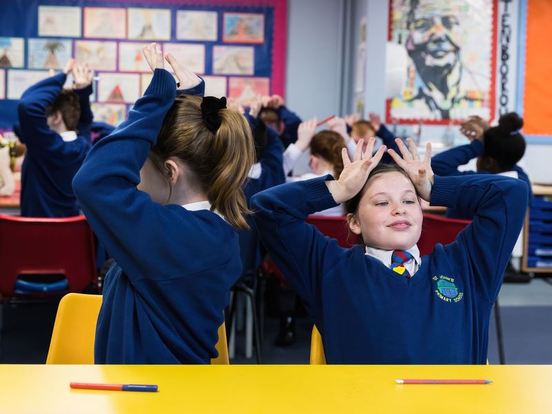 Children in a classroom wearing blue school jumpers and putting their hands up on their heads to resemble dog ears