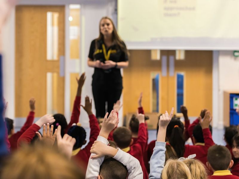 An education officer standing in front of lots of children sitting on the floor in an assembly hall, with their hands up