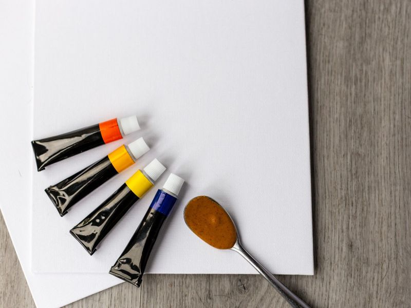 an image of a white canvas, 4 paint tubs and a teaspoon with some peanut butter on it