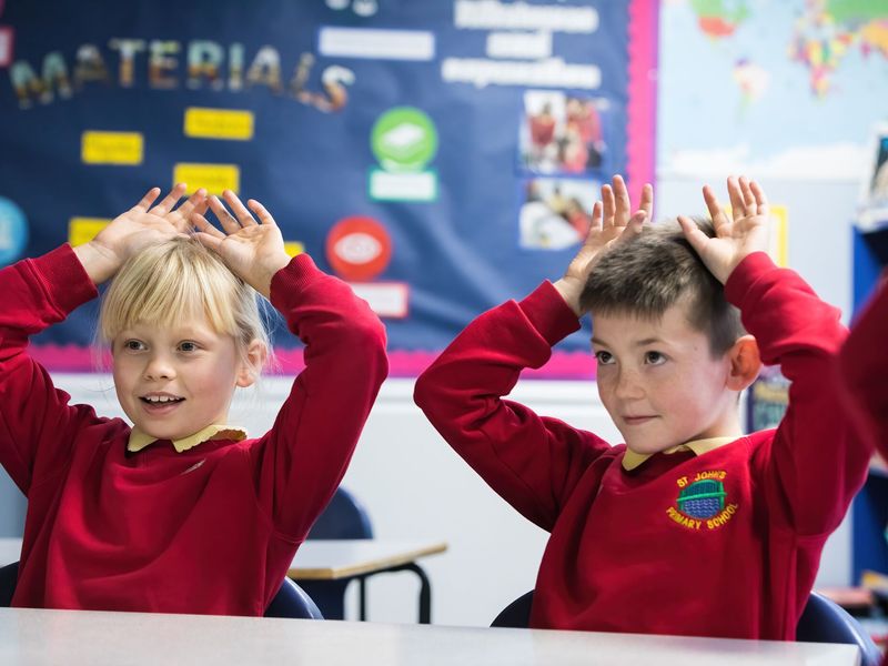Two children, a boy and a girl, putting their hands above their heads to look like dog ears, wearing red jumpers in a classroom