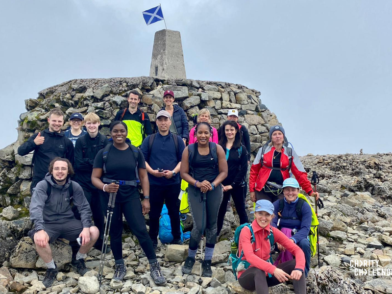 a group of people in hiking gear standing in front of Ben Nevis mountain's top smiling, with a Scottish flag on it's summit