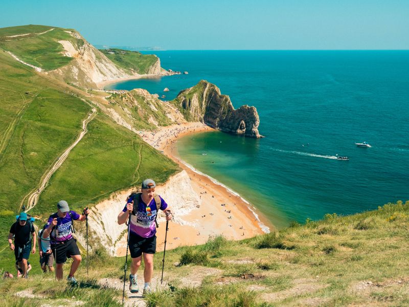 a line of people with hiking sticks walking up a green hill along the jurassic coast, with blue skies and ocean in the background