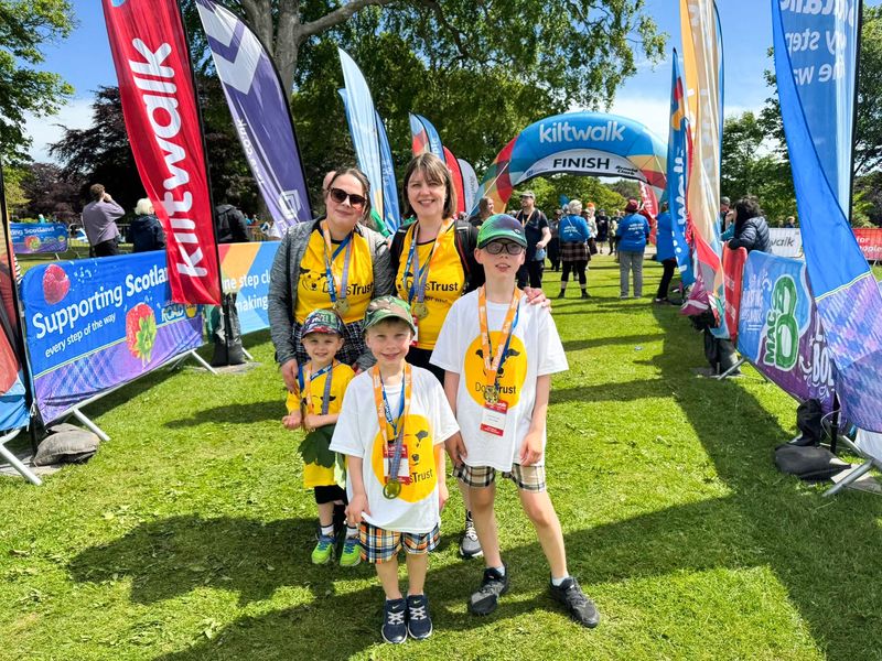 two ladies and 3 small boys smiling and wearing dogs trust tshirts and medals, standing in front of a race finish line