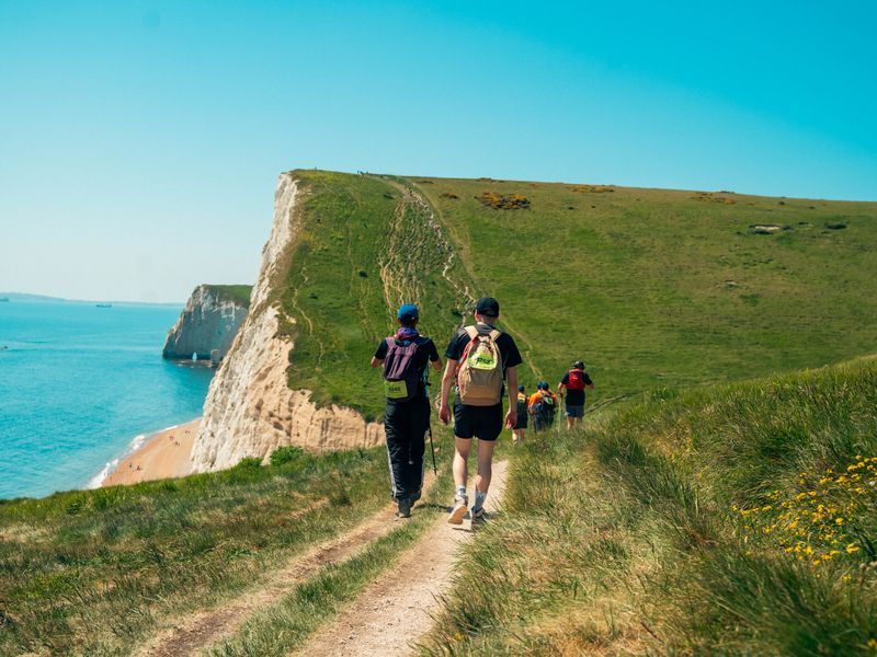a line of people with hiking sticks walking up a green hill along the jurassic coast, with blue skies and ocean in the background