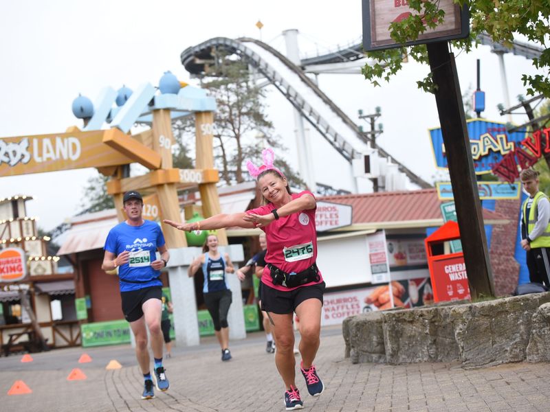A lady posing for the camera with a man running behind her, whilst jogging around Thorpe them park