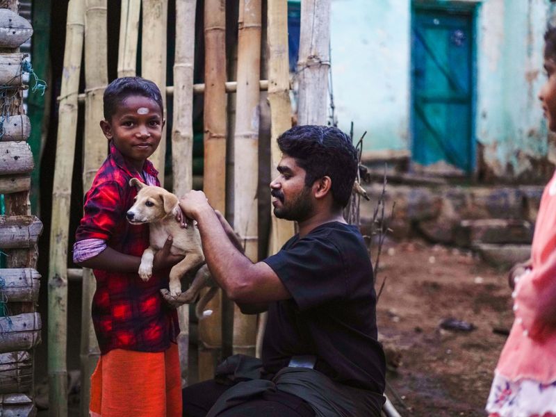 A child holds a small light‑brown puppy while an adult kneels to gently examine it in a rural outdoor setting beside a bamboo fence and a blue‑doored building.