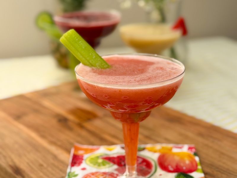 A pink frothy drink in a cocktail glass with a celery garnish, with two other colourful drinks blurred in the background.