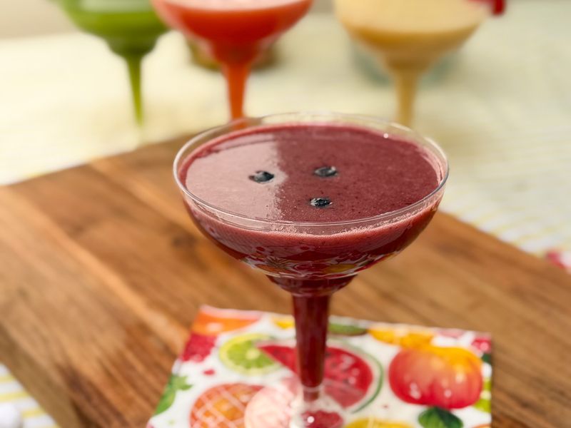 A purple smoothie in a glass with berries on top, with other colourful smoothies blurred behind.