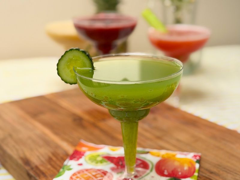 A green cocktail in a stemmed glass with a cucumber slice on the rim, set on a colourful fruit-patterned napkin, with three blurred cocktails in red, yellow and pink behind.