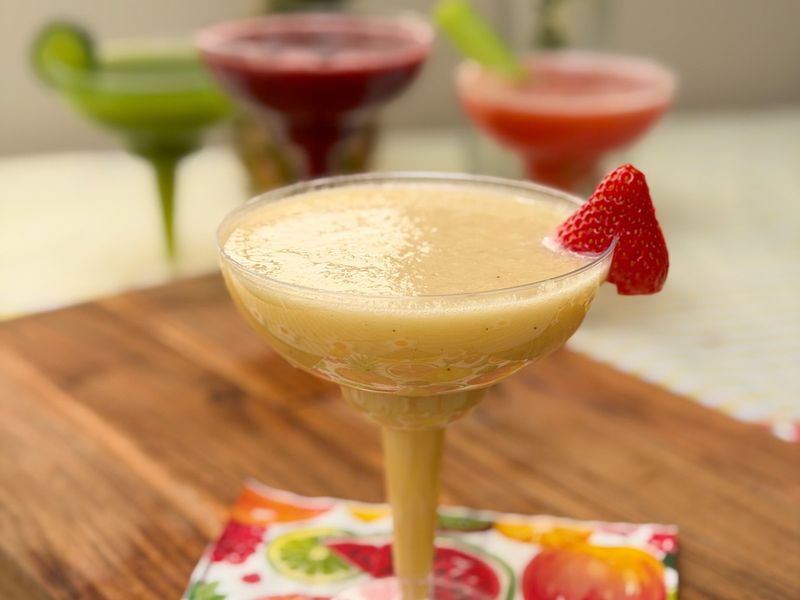 A yellow frozen drink with a strawberry garnish, with three colourful drinks behind it.